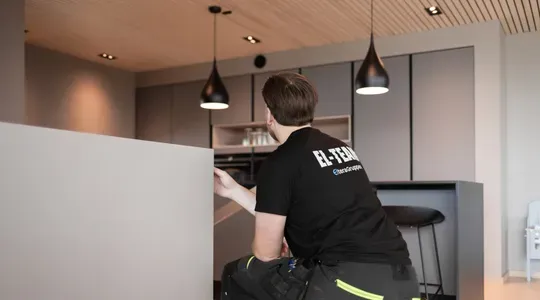 En elektriker i svart t-skjorte arbeider i et moderne kjøkken med grå innredning og hengende lamper.