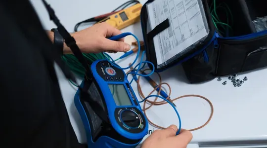 En person bruker et blått måleinstrument ved et bord, med ulike ledninger og et åpent utstyrsett med dokumenter.