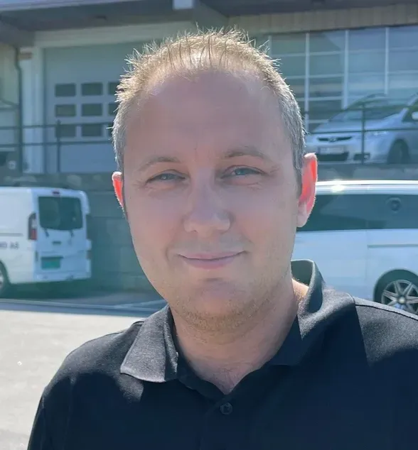 En mann med kort hår i sort poloskjorte står utendørs foran et industribygg og noen varebiler.