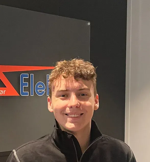 Ung mann med kort, krøllete hår smiler og ser inn i kameraet. Han har på seg mørk arbeidsjakke med logoen "Strøm Elektro". Bakgrunnen er et kontor med mørke vegger og et skilt med firmalogo.