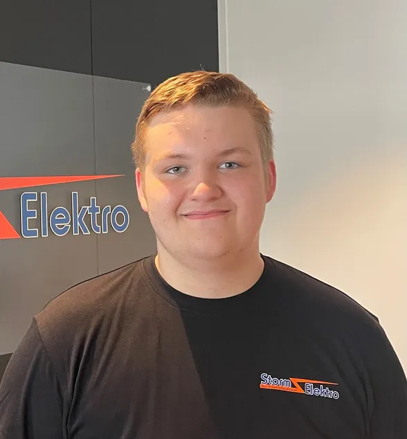 Ung mann med kort lyst hår smiler foran en vegg med «Elektro»-skilt. Han har på seg en svart t-skjorte med firmalogoen «Strøm Elektro».
