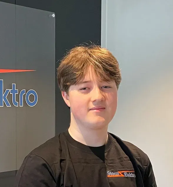 Ung person med brunt hår står foran et skilt med teksten 'Elektro'. Personen har på svart t-skjorte med logoen 'Storm Elektro'.