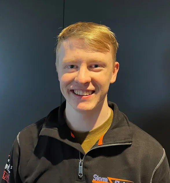 Ung mann med lyst hår smiler foran en mørk bakgrunn. Han har på seg en svart jakke med logoen 'Stom Elektro'.