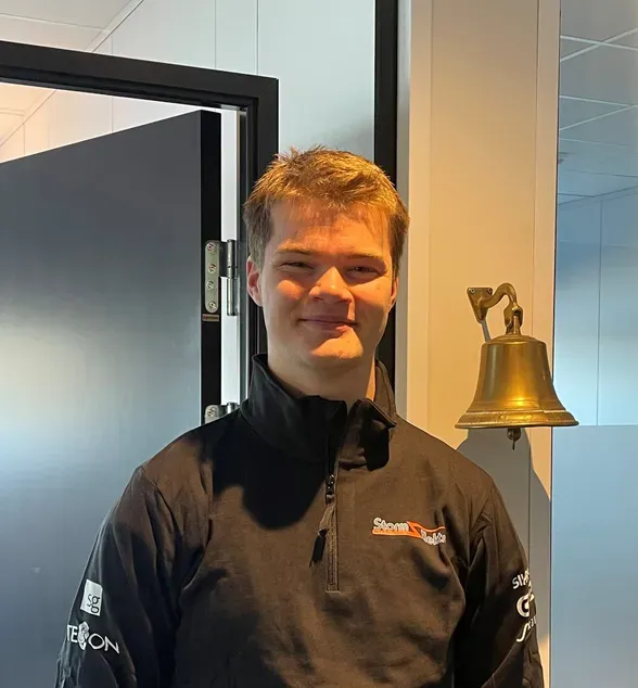 Ung mann med kort hår står innendørs ved siden av en messingklokke, iført sort jakke med firmalogo.