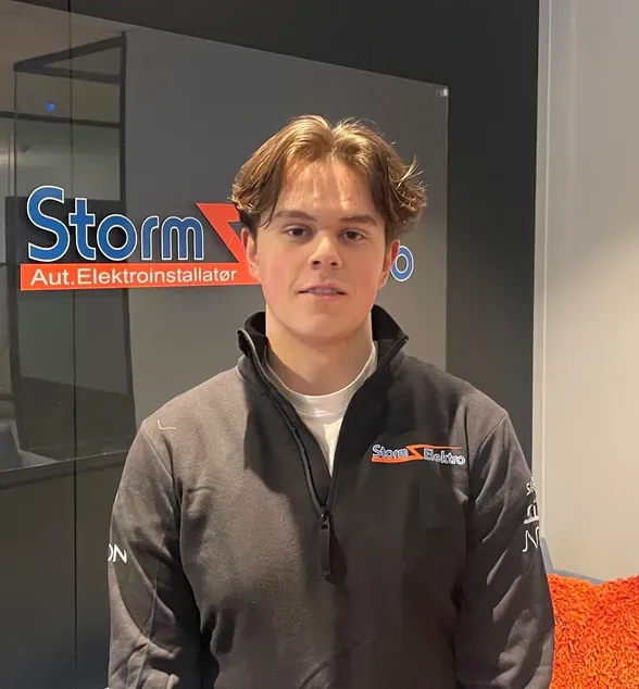 En ung mann med brunt hår står foran en vegg med logo og teksten 'Storm Aut. Elektroinstallatør'. Han har på seg en mørk genser med samme logo.
