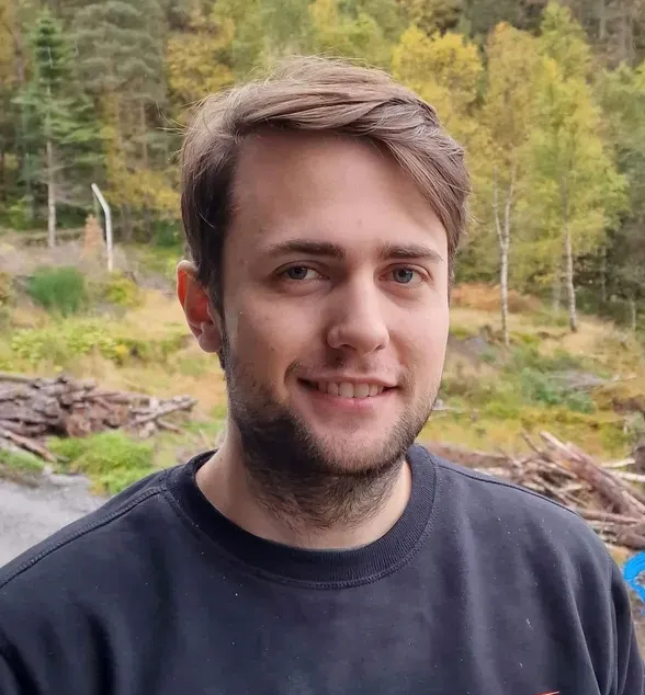 Ung mann med mørkt hår og skjegg, iført en mørk genser med logoen 'Storm Elektro', står utendørs foran et område med trær og vedstabler.