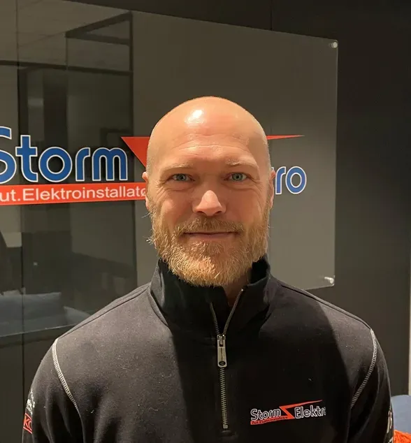 En mann med skjegg og barbert hode står foran en vegg med firmalogo. Han har på seg en svart genser med logoen til Storm Elektro.