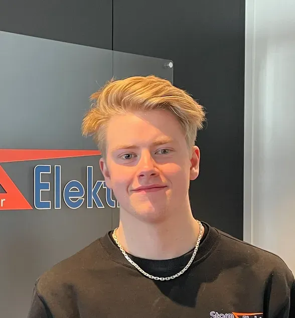 Ung mann med lyst hår, kledd i en svart genser med logoen til Storm Elektro, står foran et skilt med samme logo.