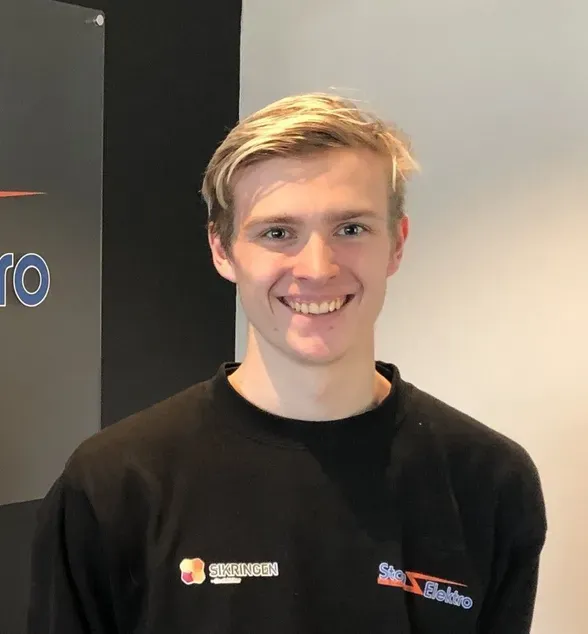Ung mann med lyst hår smiler og ser mot kamera. Han har på seg en sort genser med bedriftslogo og står innendørs foran en lysegrå vegg.