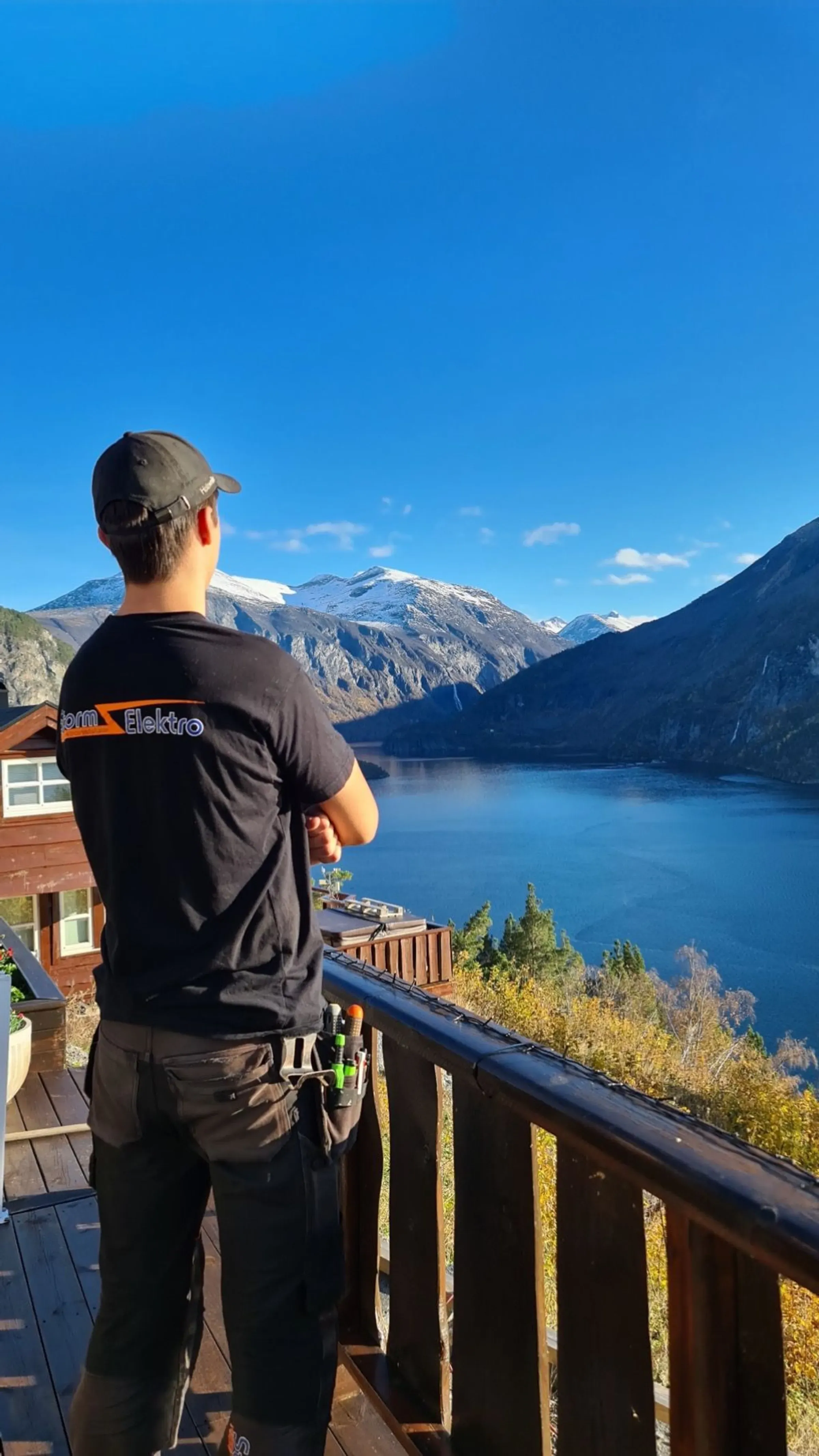 En mann står på en terrasse og ser utover en vakker fjord omgitt av fjell og natur under en klar blå himmel.