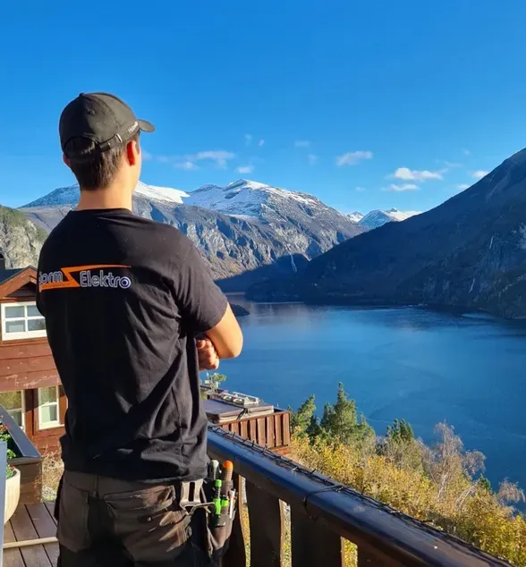 En mann står på en terrasse og ser utover en vakker fjord omgitt av fjell og natur under en klar blå himmel.