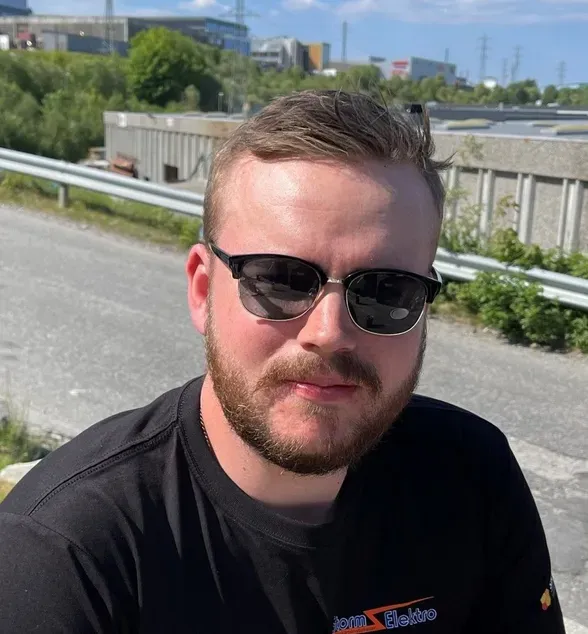 En mann med solbriller og skjegg sitter utendørs ved en vei på en solrik dag.