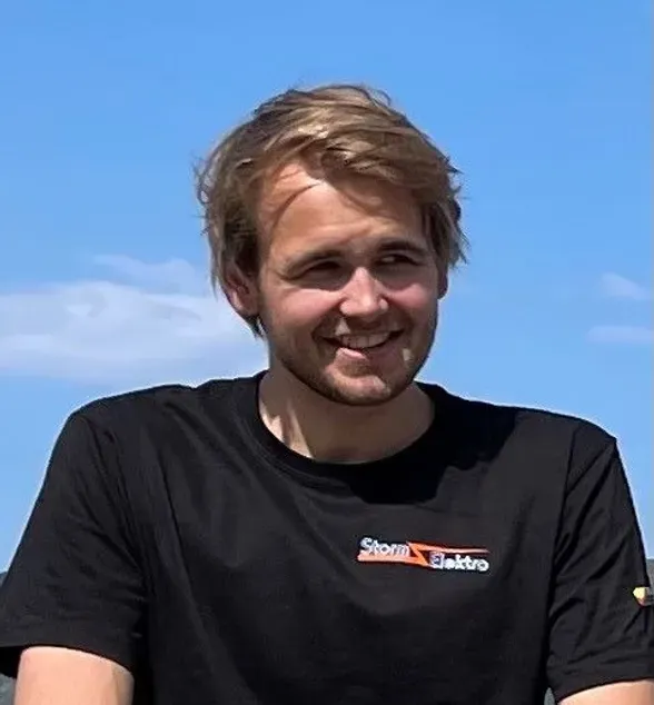 En ung mann med lyst hår smiler foran en blå himmel. Han har på seg en sort t-skjorte med logoen 'Storm Elektro'.