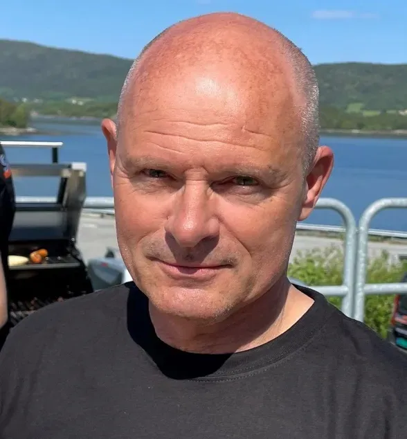 Skallet mann med svart t-skjorte, står utendørs ved vannet med grill og fjell i bakgrunnen.