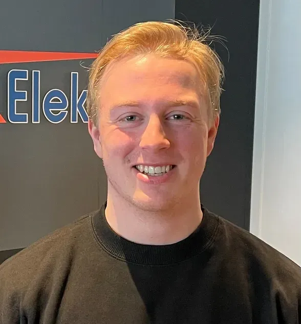 En smilende ung mann med lyst hår står foran et skilt som sier "Storm Elektro". Han har på seg en sort genser med logoer.