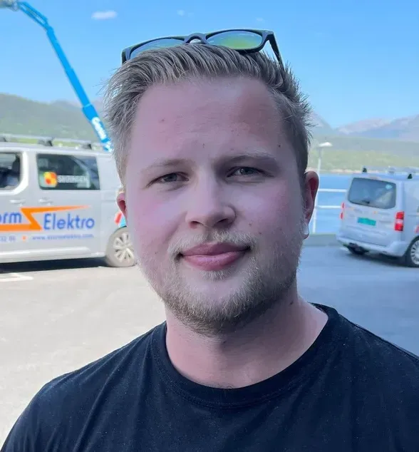 En ung mann med blondt hår, iført sort t-skjorte med logoen «Storm Elektro», står ute foran varebiler med samme firmalogo. Han har solbriller på hodet og det er fjell og blå himmel i bakgrunnen.