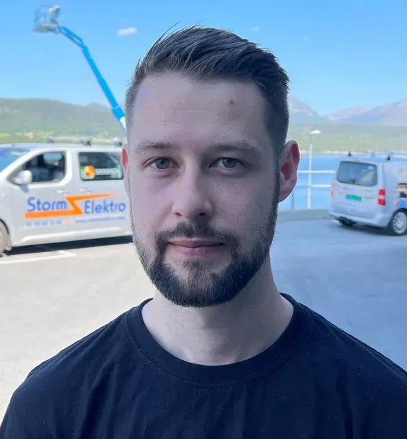 En mann med kort brunt hår og skjegg står utendørs. Han har på seg en svart t-skjorte med Storm Elektro-logoen. I bakgrunnen står to firmabiler og en person i lift, med fjell og blå himmel bak.