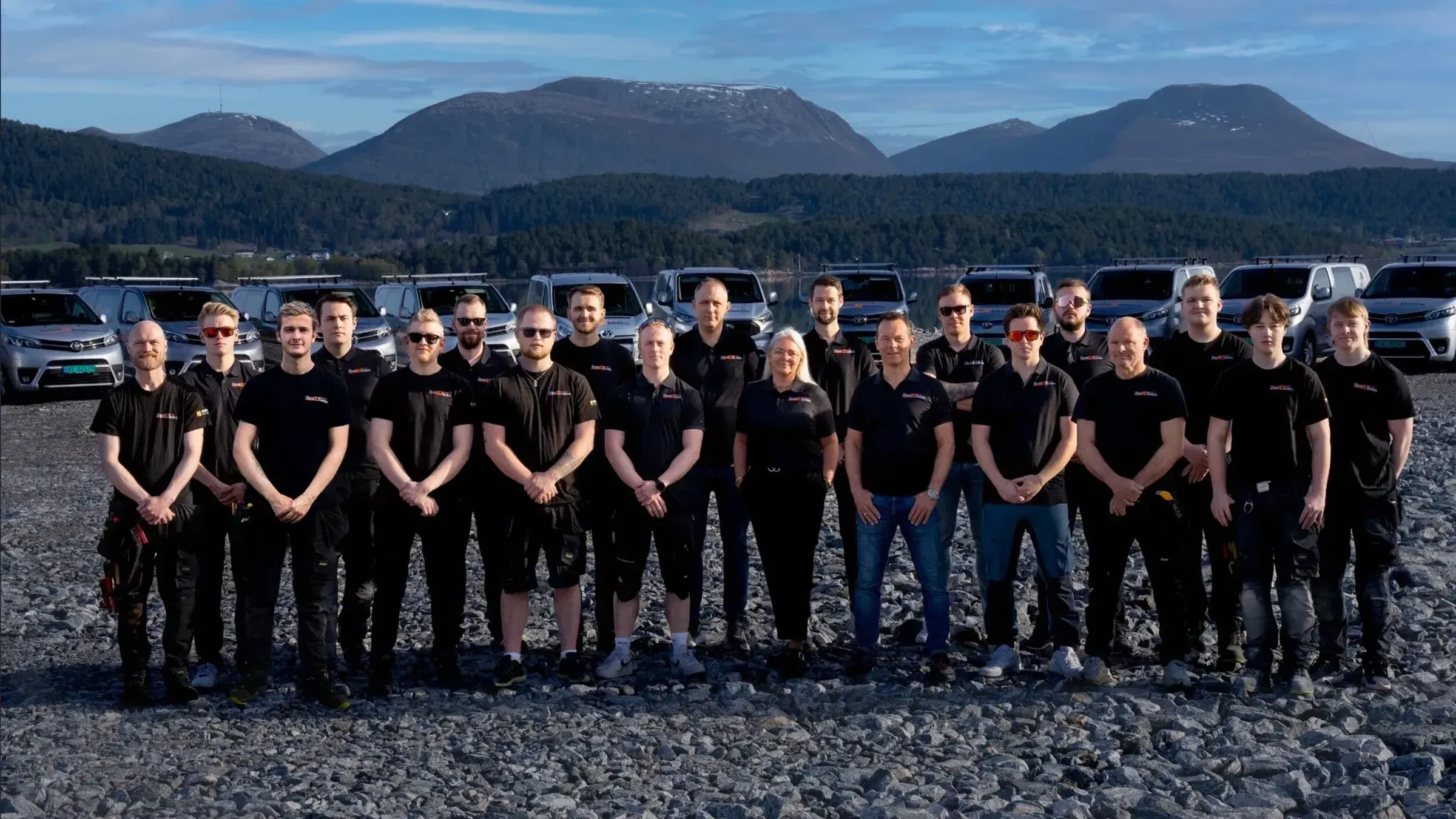 En gruppe mennesker står ute på en grusplass foran flere parkerte biler, med fjell og blå himmel i bakgrunnen.