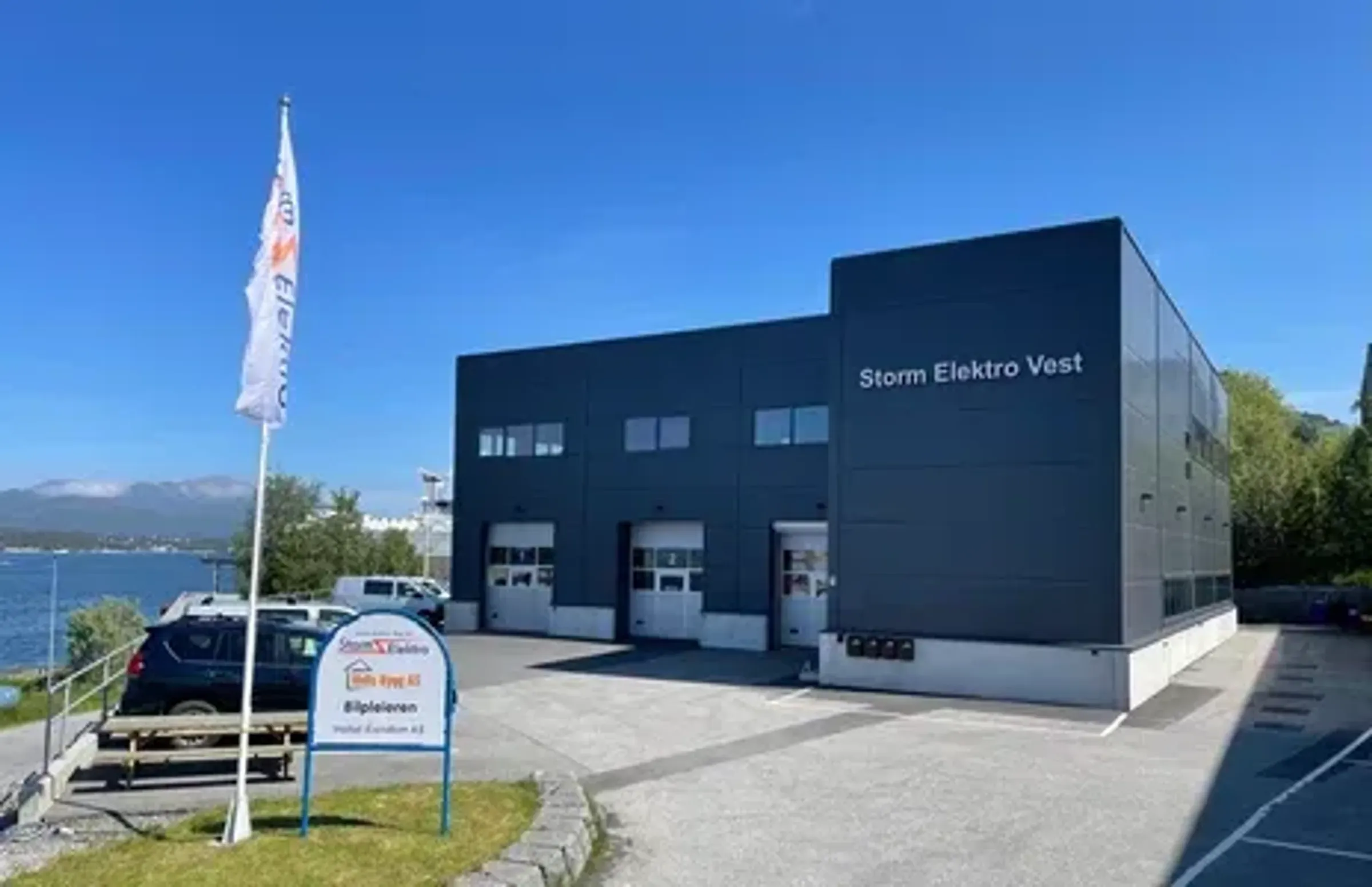 En moderne, mørk grå industribygning merket 'Storm Elektro Vest' sett fra utsiden, med flagg og skilt foran bygget. Ligger ved vannkanten, under en klar blå himmel.