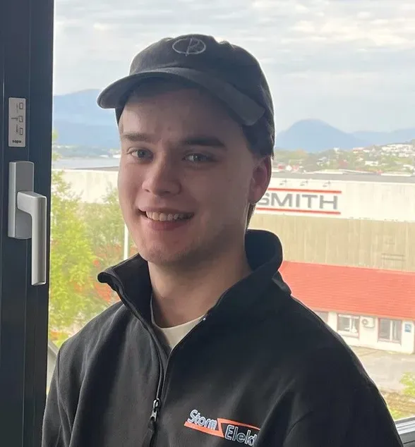 Ung mann med mørk caps og jakke smiler foran et vindu med utsikt mot fjord og fjell.